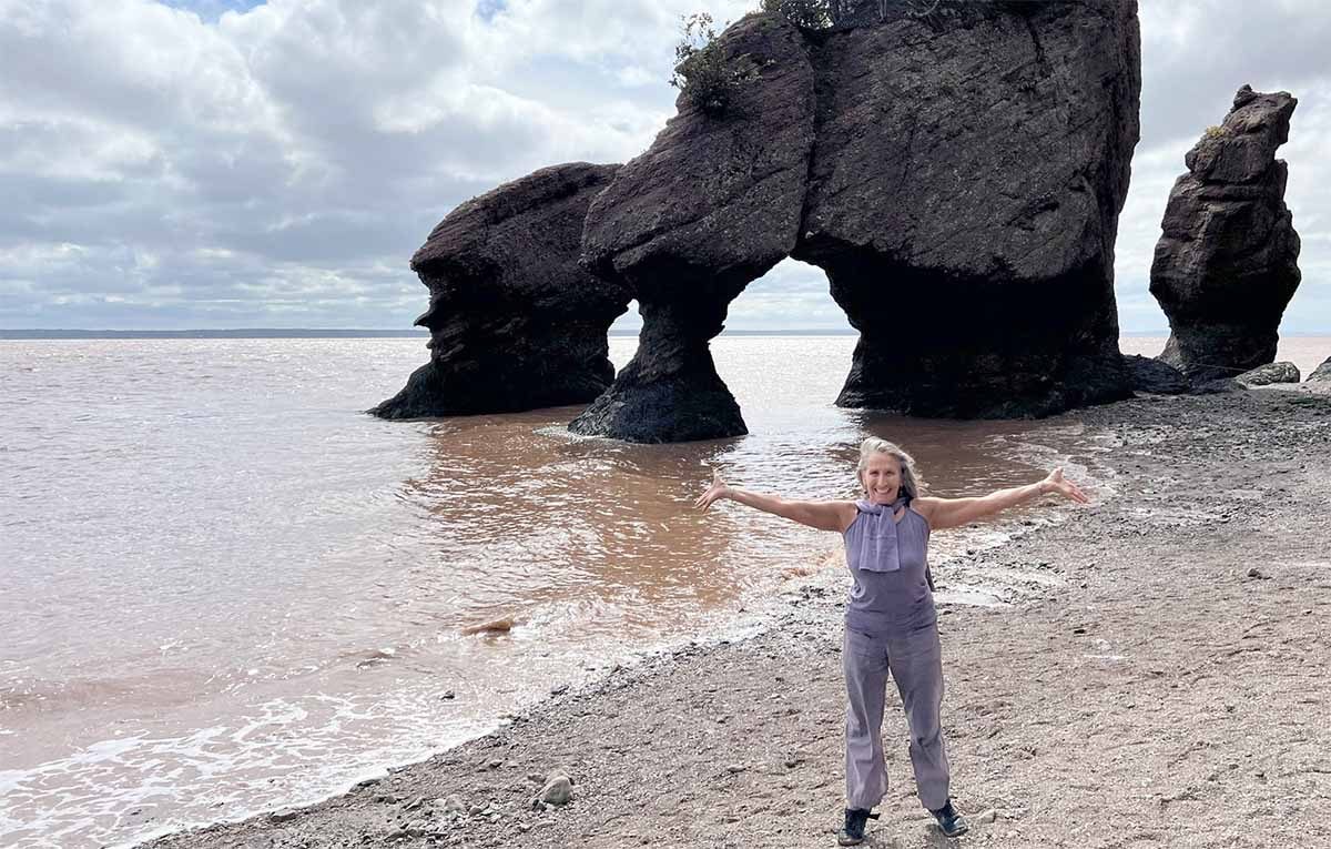 Ajeet Khalsa with arms outstretched on a beach beside a rock arch
